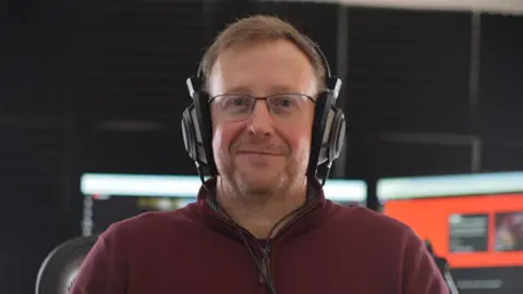 Jamie Niblock/BBC TheRunningManZ sits in a gaming chair with headphones on as he smiles at the camera. Computer screens that are switched on and blurred can be seen behind him.