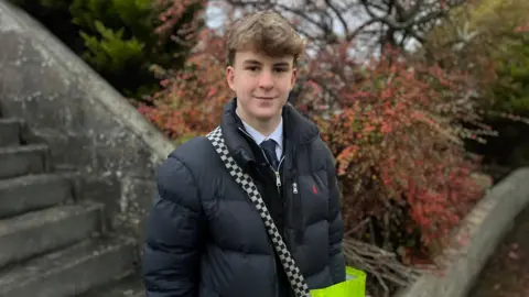 Harry, a good-looking teenager, with a modern hairstyle, wears a black puffer jacket, black cardigan and school shirt and tie underneath. He stands outside a property, carrying his large papers bag - a hi-vis yellow thing.