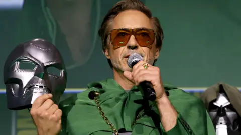 Getty Images Robert Downey Jr at San Diego's Comic Con event in 2024, when his return to the MCU in a new role was announced on stage. He is wearing the character's recognisable shade of dark green and sunglasses, having just removed Doom's metallic mask, which was hiding his identity from the audience, which he is now addressing using a microphone.