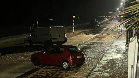 Claire Gear A number of cars appear randomly scattered across a beach front road, which is very wet. It is dark. 