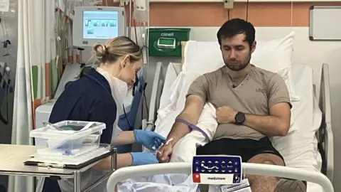 BBC Jordan lying on a bed during a stem cell donation session at the Anthony Nolan Cell Collection centre in Nottingham. A nurse is sat alongside him carrying out some medical checks a nearby machine extracts stem cells from his blood, 