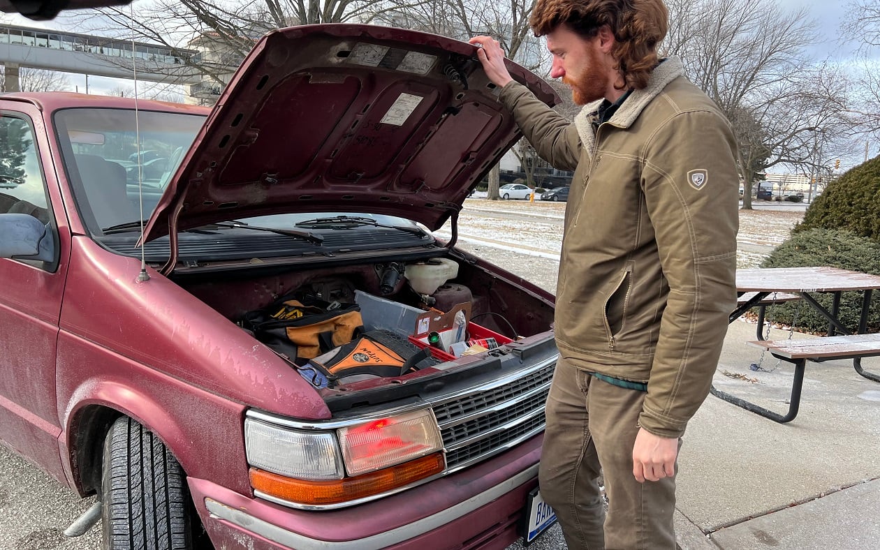 Man stands with a trunk open that has tools in it - in an old red vehicle