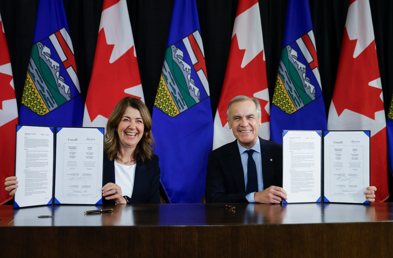 two smiling politicians hold up folder showing signed agreement, Alberta and Canada flags behind them