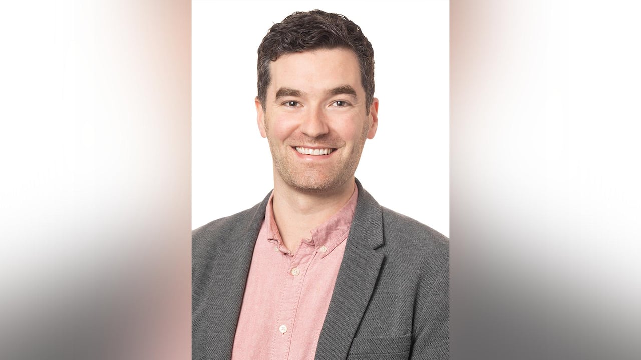 A portrait of a smiling man wearing a dusty pink button down dress shirt and grey blazer.