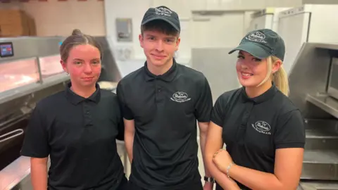 BBC Two young women and one young man (centre) in their chip shop uniforms of a black polo shirt and trousers and a skip cap smile for the camera. They are standing beside the fat fryers and food prep area, all in stainless steel.