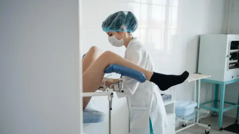 Getty Images A female medical professional wearing scrubs, a face mask and a hair net performing a cervical cancer screening on a patient. Only the legs of the patient can be seen from a side angle. 