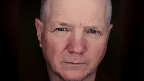 Haemochromatosis UK Finbar Polin is staring intently at the camera. It is a close up of his face against a black background. He has short grey hair and blue eyes.