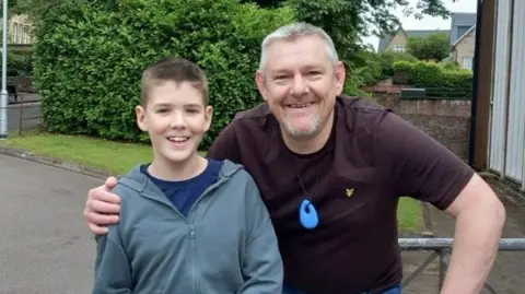 Sharon Knight There is a young boy wearing a grey coat and blue pants. He has brown hair. He is being hugged by an older man with grey hair. He is wearing a maroon t-shirt, around his neck is a blue silicone toy. They are stood in front of some trees. They are both smiling. 