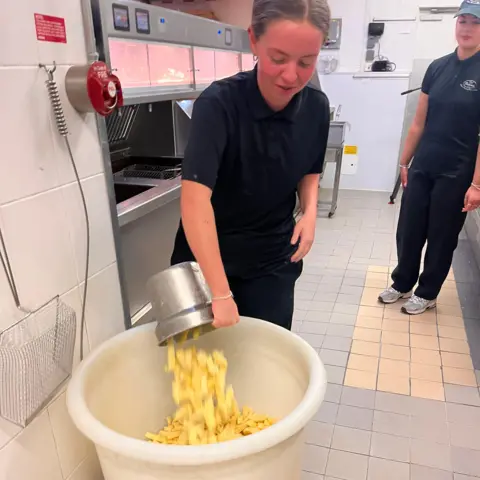 Kenzie pours chips from a stainless steel container into a large white plastic bucket in the chip shop. The chip fryers and hot food display cases are behind her facing the customer area.