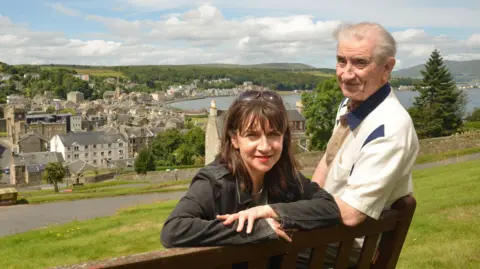 Shutterstock Maureen Beattie and her late father Johnny Beattie in an old photo leaning on a park bench in Rothesay, Isle of Bute. They are on top of a hill overlooking the town and the water below.