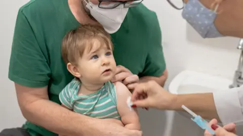 Getty Images A young toddler looks up at a doctor who is holding a vaccine while rubbing a piece of cotton wool on his arm. The toddler is wearing a green and white striped T-shirt and is sitting on his father's knee, who is also wearing a green top.