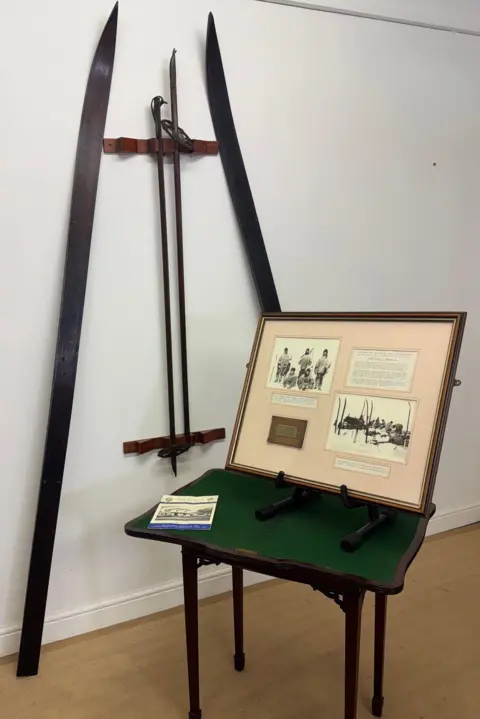 Adam Partridge Auctioneers The pair of skis and ski poles are mounted on a white wall and there is a presentation case open which features two group black and white photos, a brass plaque and annotations relating to the expedition.
