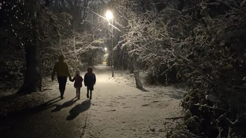 Rachel de Kam  An adult and two young children walking on a snowy street at night-time