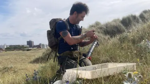 Vicki Richardson A man in his forties wearing a backpack sits in a field smiling. He has a plastic container and a plastic cylinder next to him and he is examining something in his hand. 