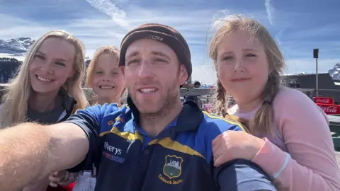 James Rogers/PA James holds out the camera for a selfie with his family of two girls and his partner Ellie. They appear to be in a ski resort as there are snowy mountain tops behind. He wears short sleeved T-shirt and beany hat. 