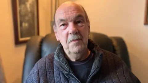 BBC Anton Hammer, 72 wearing a dark top with a balding head and dark eyes. He is sitting in an armchair and smiling into the camera.