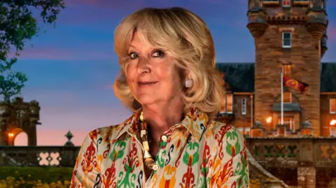 BBC/PA Handout photo of Fiona, one of the contestants in series four of BBC1's The Traitors. Fiona has short blown curled hair, blue eyes and smiles at the camera. She wears a yellow, green and red patterned blouse, a thick shell necklace and diamond earrings. A UGC image of the castle is seen in the background. 