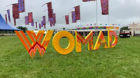 BBC Womad sign at the festival grounds. The abbreviation is spelled out with large, orange and yellow printed letters on grass. The festival site can be seen behind in the distance - there are colourful flags, tents and stands. 