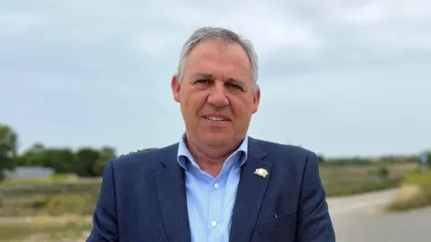 Constable Andy Jehan, a man with short grey hair. He is clean shaven, and wearing a navy blue blazer over a sky blue shirt with white buttons. His top button is unbuttoned. He has a badge on his left lapel. Behind him, out of focus, is a road and green fields.