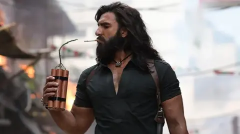 Jio Studios Ranveer Singh in a still from Dhurandhar, seen holding dynamite to light a cigarette. He's wearing a black shirt with a silver pendant around his neck. He has long hair and a beard.