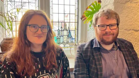 BBC A woman with long ginger hair, black framed glasses and a blue patterned jumper next to a man with ginger hair, a ginger beard and black framed glasses in a blue shirt and blue chequed jacket in front of a stained glass window