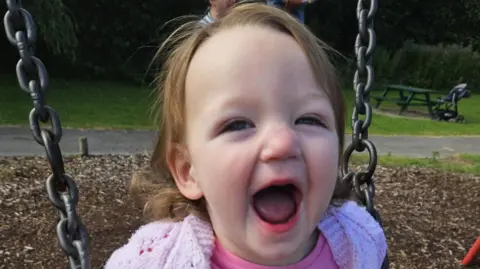 Story family A head and shoulders picture of a two-year-old girl while on a swing in a children's park. She has short brown hair and is smiling broadly at the camera. She is wearing a pink cardigan and pink top.