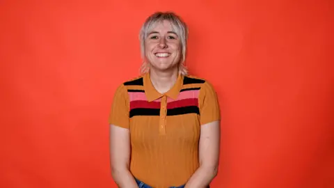 BBC Maisie Adams wearing a yellow striped top with pink and red on it. She has blonde short hair and is standing in front of a red background. 