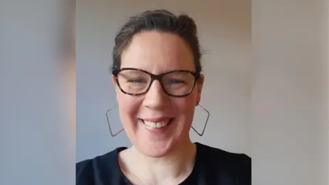 Supplied Lucy is looking at the camera and can be seen from the shoulders up. She's wearing large square-hooped earrings and smiling, with her hair back.