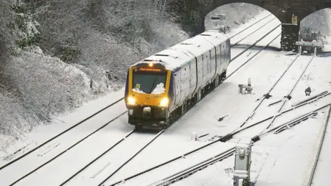 PA Media A wide shot of a yellow train travelling through snow. The track is covered with snow, and the train is covered with snow.