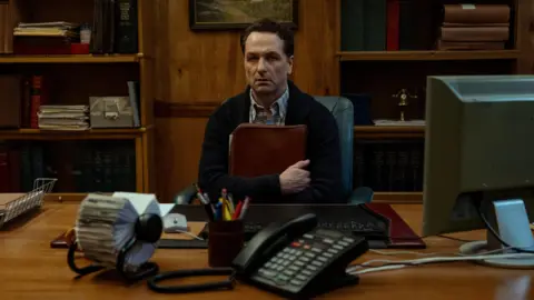 Apple TV Actor Matthew Rhys sits in a chair at a wooden desk holding a folder to his chest. On the table is a black landline telephone, a rolodex, a computer and a pot of pens. He stares into the distance with a grave look on his face. 