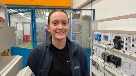 BBC A young woman is wearing a Siemens branded blue fleece and is standing in a room surrounded by electrical equipment