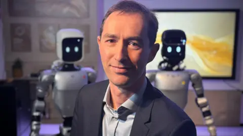 BBC Tim Ensor is wearing a blue suit and shirt. He is smiling at the camera and has a robot looking over each of his shoulders. The robots are humanoids - so have a head, body, arms and legs. Their heads are cubes with black screens for faces, displaying blinking white oval eyes. 