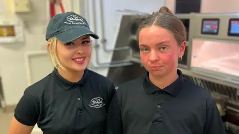 16-year-old Erin and 18-year-old Kenzie stand side-by-side in the Ashvale chip shop, the big fryers behind them. They both wear a black uniform and hat.
