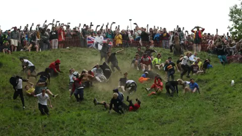 Reuters A group of men are hurling themselves down a steep hill, some are wearing fancy dress and others are wearing T-shirts and jogging bottoms. At the top are a big group of spectators pointing their phones downwards.