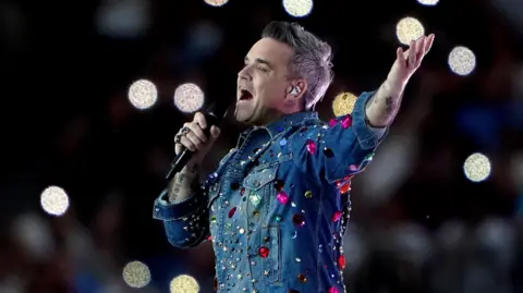 PA Media Robbie Williams wearing a denim shirt with jewels stuck on it. He is pictured side-on while performing at a concert. He is singing into a microphone held in his right hand while his left arm is raised in the air.