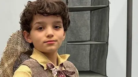 Contributed Tyne, wearing a Munchkin costume, looking at the camera. He has short dark hair, has a hat on his back, a yellow shirt, tie round his neck, and brown jacket. A rail is to his left. 