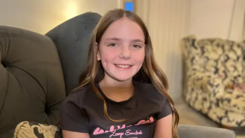 BBC A young girl with long brown hair smiles into the camera. She is wearing a grey t-shirt with bold pink writing across it. 