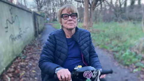 Elizabeth Baines / BBC A woman sits on an electric scooter. She has large black glasses and short blonde hair. Behind her, the Meanwood Valley Trail is blurred.
