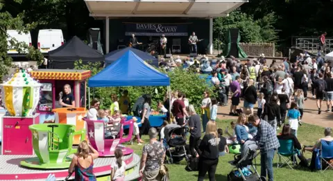 Keynsham Music Festival A small summer festival on a sunny day. Crowds of people including parents with prams sit or stand on a patch of grass in front of a stage where a band is performing. A spinning teacup ride is visible in the foreground.  