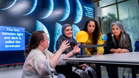 University of Leeds/Mark Bickerdike Four people are in a classroom. Three are sat at a table, one is crouched down next to them. On the table are models of the solar system. There is a screen behind them with images of the moon on.