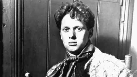Getty Images/Hulton Archive A black and white photo of a young Dylan Thomas, wearing a cravat and looking straight at the camera. He is sitting down and has short curly hair. 