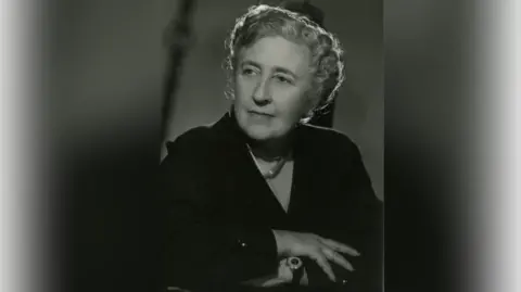 Christie Archive Trust An archive sepia image of Agatha Christie, around 1950. She is sitting down with her arms folded and looking away from the camera.