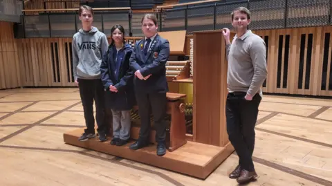 BBC Three teenagers and a man are standing in front of an organ on a stage, looking at the camera.