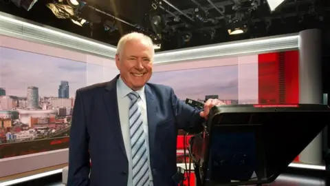 BBC BBC TV presenter Nick Owen stands in the studio leaning on one of the cameras, while smiling. The Birmingham landscape is projected on the screen behind him.