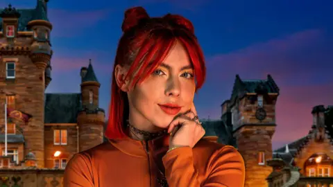 BBC/PA Jessie Stride in her promotional image for The Traitors Series 3. She is wearing an orange top and has red hair. it is a close up image showing her head, shoulders and arm. in the background there is a picture of The Traitors castle