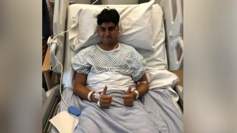 Ekam Dhaliwal A young man with dark brown hair is in a blue and white hospital gown sitting on a hospital bed. He has his thumbs up  and is smiling at the camera